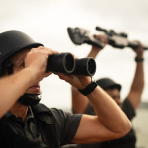 Ambrey team leader with binoculars stands lookout while second team member hold up rifle.